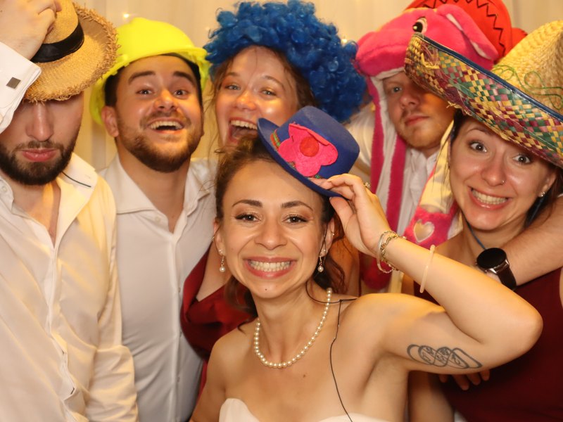 Sechs Hochzeitsgäste lachen mit bunten Hüten und Perücken in der Fotobox.